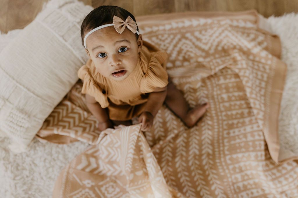 adorable little girl wearing a boho brown dress with a brown ribbon headband looking up at the camera sitting on our dusty pink swaddle receiving blankets