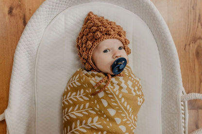 cute little baby wearing a crochet brown bonnet wrapped in our dark yellow blanket lying in a cotton-lined white baby bassinet 