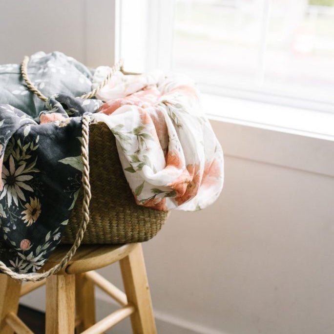 peony cotton swaddle and spring blossom blanket tossed inside a woven basket placed on top of a chair near the light and airy window 