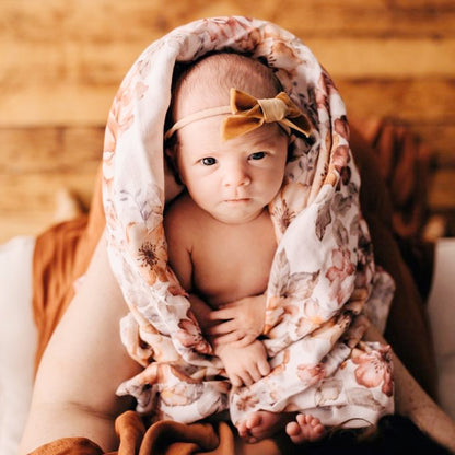 Baby in a swaddle surrounded by primroses and peonies in vintage colors. Baby is dressed cutely in a light brown bow and is looking up at mama.