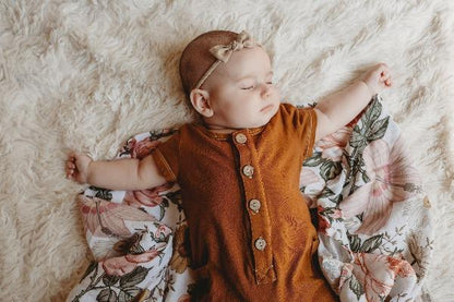 A baby girl is sleeping soundly, arms out, eyes closed on a soft baby girl swaddle blanket from the mini wander. She is wearing a burnt orange colored bodysuit and a beige bow headband. The cotton blanket is covered in pretty garden flowers.