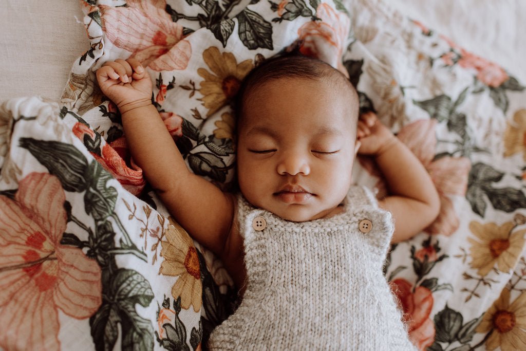 A tiny baby girl with squishy cheeks is taking a nap with a floral lovey. The lovey is a soft muslin swaddling blanket covered in flowers. Her arms are up and she is wearing knitted overalls.
