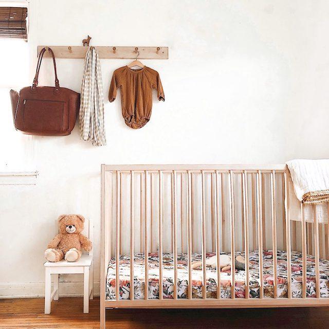 boho nursery inspiration exhibiting the classic and timeless vintage floral crib sheet inside the crib adorned with a brown bear on a chair, a floating wooden hanger rack with brown top and a bag