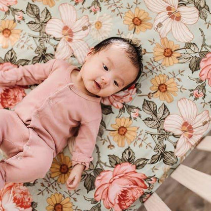 Baby is dressed in blush pink onesie and is sleeping in a cribsheet with a floral design on a green background. Daisies, roses, and creamy pink hibiscus are among the flowers printed on the cribsheet.