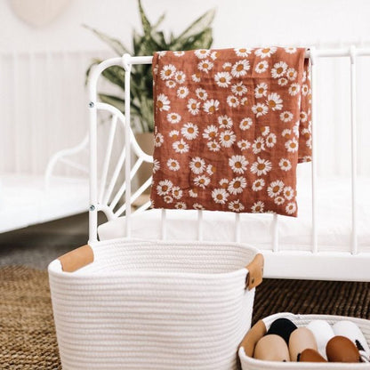 In a white-boho-themed bedroom, an earthy clay brown swaddle with Daisy floral design hangs on the bed rail.