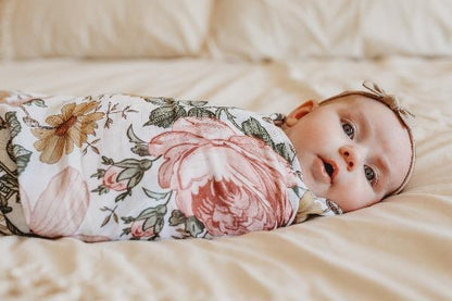 A sweet baby girl is laying down, swaddled in a wrap that has large roses, green leaves and yellow daisy flowers. The swaddle blanket is from the mini scout brand, now known as Mini Wander.