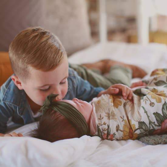 A newborn family photoshoot. A baby girl is swaddled in a floral wrap with a green bow, her brother is kissing her forehead.