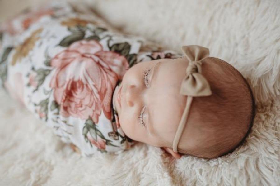 a photo of a sleeping baby girl swaddled in a vintage floral muslin blanket. she has a khaki colored petal bow on her head.