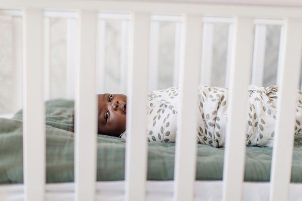 little one wrapped in the leaves sage green summer swaddlepeeking into the camera from inside his crib 