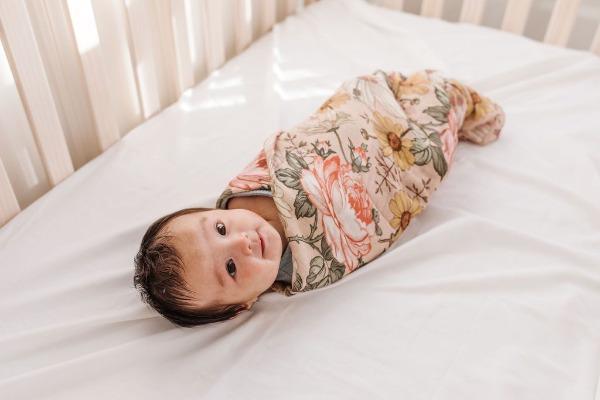 A happy baby lies in his crib, wrapped in a rose pink baby swaddle with garden floral designs such as hibiscus, rose, and daisy.