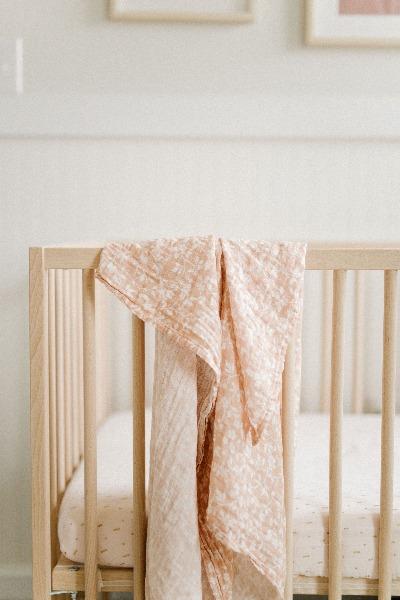pink cotton swaddle blanket with magnolias hanging on the side of the wooden crib