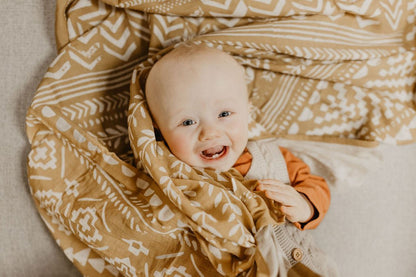 a smiling one-toothed baby wearing neutral clothing looking at the camera with our boho dark yellow blanket draped all over him towards his back