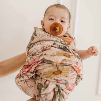 Baby is held in someone's arms and wrapped in a rose pink baby swaddle with hibiscus, rose, and daisy garden floral design.
