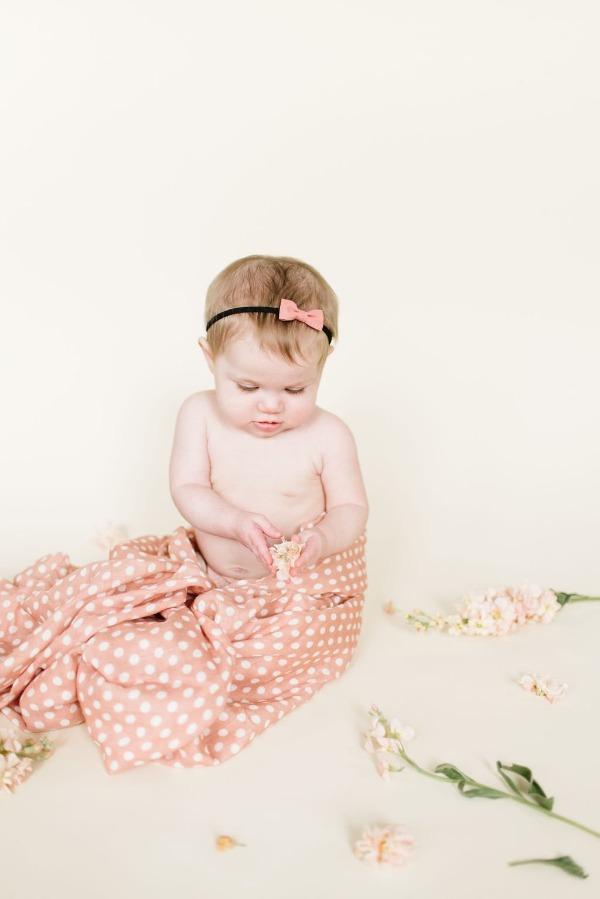 adorable baby girl with pink ribbon headband playing with flowers and petals half wrapped in the blush pink swaddle blanket