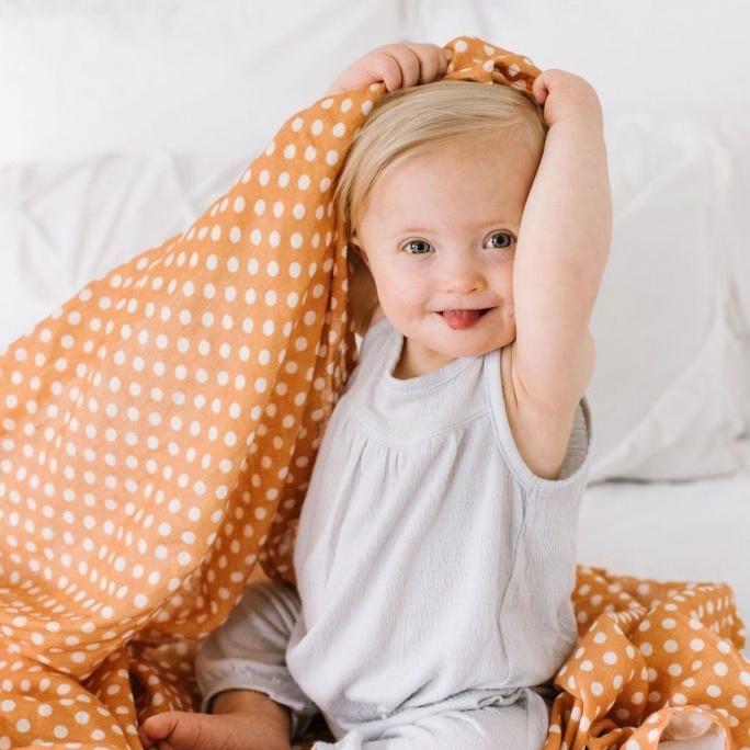 adorable baby girl with blonde hair playing with the amber polka dot swaddle blanket placing it on her head