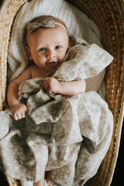 a sweet smiling baby girl wearing a headband inside her baby basket hugging her floral gray baby blanket in the magnolia tree print