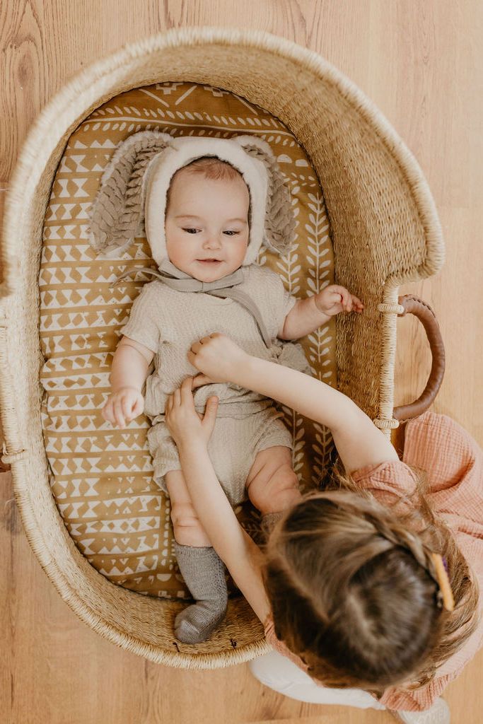 photo of two adorable babies showing a little baby wearing a cute animal hat and neutral baby clothes lying on a baby bassinet lined with our dark yellow blanket while the big sister in a pink dress fixing baby's clothes